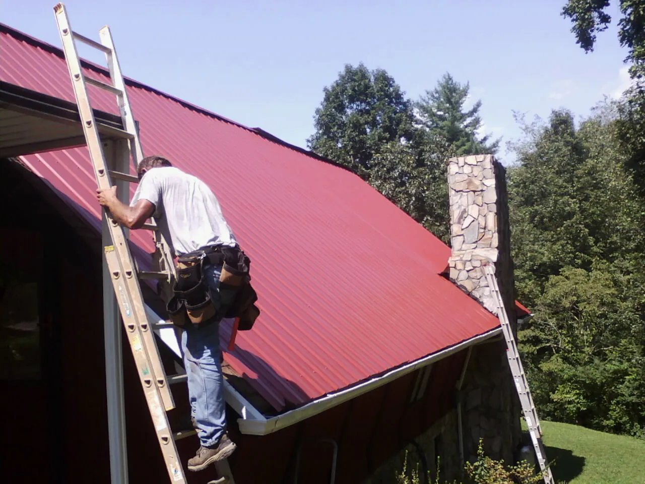 Roofer on a ladder beside a red metal roof and stone chimney — Soffit & Fascia Repair by Roofing Brothers in New Straitsville